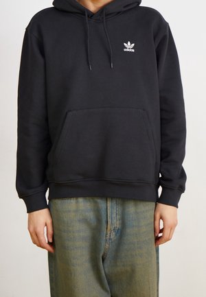 Osoba w czarnej bluzie Adidas z kapturem, z kieszenią z przodu i logo, w połączeniu z luźnymi, wyblakłymi niebieskimi dżinsami.