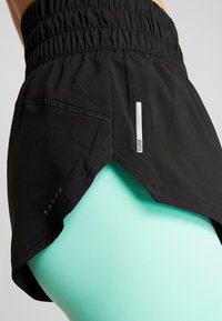 Black athletic skirt with a gathered waist, side slit, and reflective logo detail. Paired with fitted, mint green leggings underneath.