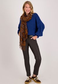 Vrouw draagt een blauwe trui, een bruine geruite sjaal, zwarte broek en bruine sneakers, staat met handen in de zakken en glimlacht.