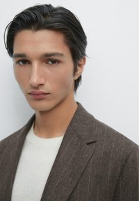Brauner Nadelstreifen-Blazer über einem hellen Pullover, Modell mit glattem schwarzen Haar, neutraler Hintergrund, Fokus auf die Gesichtszüge und Details der Kleidung.