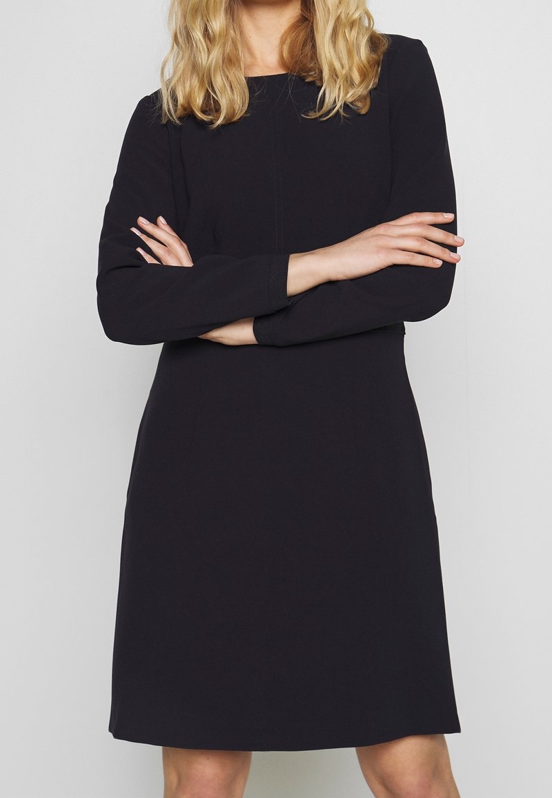 Femme blonde portant une robe noire à manches longues, les bras croisés, devant un fond clair.