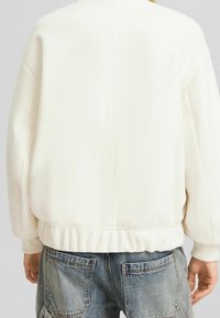 Biały, oversize'owy sweatshirt z ribbowanymi mankietami i dołem, o gładkiej fakturze i minimalistycznym wzornictwie. Widok z tyłu nie pokazuje widocznych elementów metalowych.