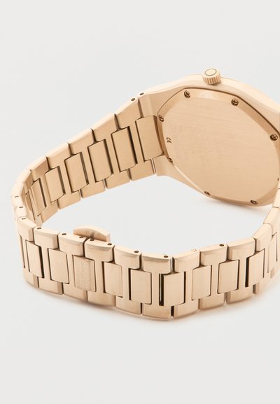 Χρυσό μεταλλικό ρολόι χειρός με συνδεδεμένο λουράκι και κλειστό κούμπωμα, εμφανιζόμενο από πίσω, τονίζοντας την κάσα του ρολογιού και το στέμμα.