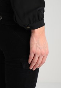 Svart blus med puffärmar och elastiska mudd, med en slät textur och metalldetalj med tryckknapp, kombinerad med svarta figursydda byxor.