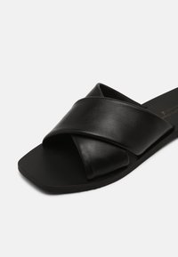 Sandale noire à glissière avec larges brides en cuir croisées et un bout carré sur fond blanc.