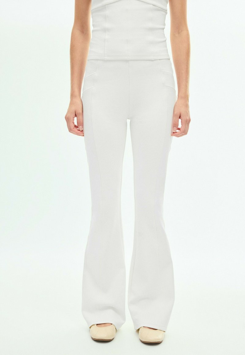 Femme portant un pantalon blanc évasé taille haute et des chaussures plates crème, debout devant un fond clair uni.