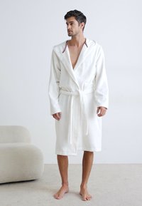 Accappatoio bianco in spugna con cappuccio, dotato di cintura legata, maniche lunghe e colletto con accenti rossi. Il modello si trova su un pavimento chiaro.