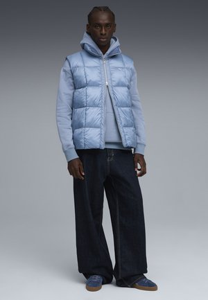 Lys blå puffervest med quiltet design over en lys blå hættetrøje, parret med mørke brede jeans og blå sneakers.