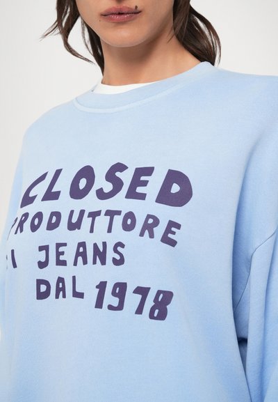 Ανοιχτό μπλε φούτερ από μαλακό βαμβάκι, με σκούρο μωβ κείμενο που αναγράφει "CLOSED PRODUTTORE JEANS DAL 1978." Απλός, με στρογγυλή λαιμόκοψη.