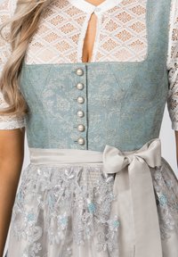Traditionelles Kleid mit einem blauen Brokat-Oberteil mit Perlenknöpfen, einer cremefarbenen Spitzenbluse und einem durchsichtigen Rock, der mit floralem Stickmuster verziert ist.