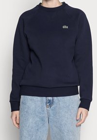 Sweat-shirt bleu marine avec un col rond côtelé et des poignets côtelés. Présente un petit logo brodé en vert et blanc sur le côté gauche de la poitrine. Texture douce.