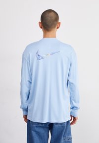 Camisa de manga longa azul claro com gola redonda, apresentando um logótipo da Nike multicolorido nas costas. Feita de um tecido de algodão suave.