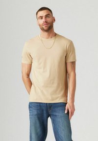 Camiseta de algodón beige con escote redondo y mangas cortas, combinada con jeans de mezclilla azules. Presenta un pequeño logotipo cerca del dobladillo.