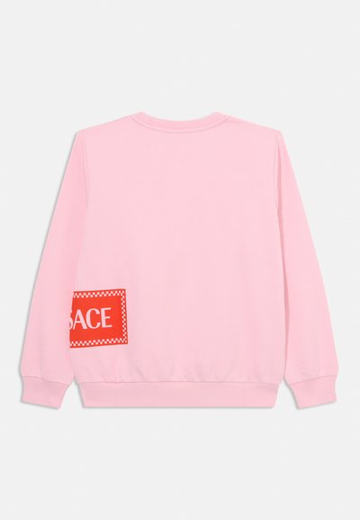 Rosa sweatshirt med långa ärmar, ribbade cuffs och en rutmönstrad röd lapp med vit text på vänster sida. Mjuk tygtextur.