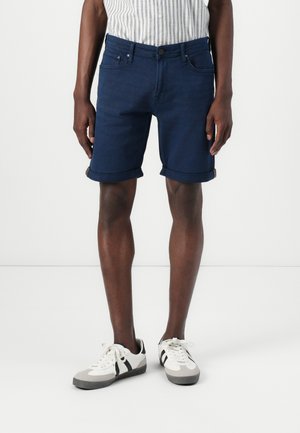 Marineblå denimshorts med op-rullet kant, fem-lomme design og subtile syninger, parret med hvide og grå sneakers.