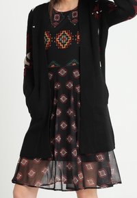 Cardigan nera lavorata a maglia con apertura frontale, caratterizzata da intricate geometrie in rosso e turchese su un vestito nero con sovrapposizione trasparente.