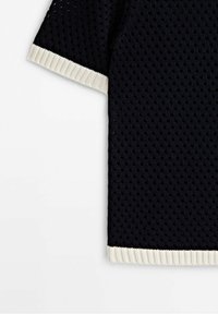 Tissu tricoté noir à manches courtes avec un tissage ouvert texturé, présentant des bords blancs côtelés aux manches et à l'ourlet.