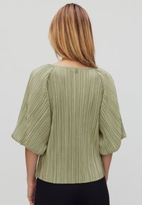 Vihreä laskostettu paita, jossa on lyhyet puffihihat ja pyöreä kaula-aukko. Takana on pieni napitusyksityiskohta. Kankaan pinta on tekstuuri.