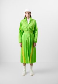 Vihreä laskostettu mekko, jossa on vetoketju edessä ja korkea kaulus, jota korostavat keltaiset yksityiskohdat; yhdistetty valkoisiin tennareihin ja sukkiin.