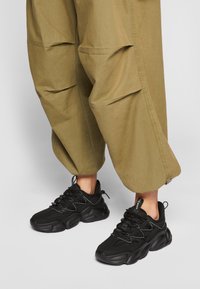 Persona vestida con pantalones holgados de color verde oliva y zapatillas negras gruesas, de pie sobre una superficie gris clara.