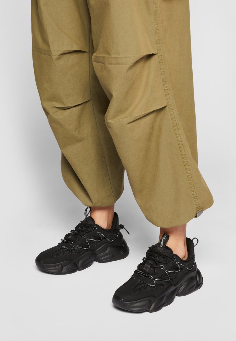 Persona che indossa pantaloni larghi verde oliva e sneaker neri spessi, in piedi su una superficie grigio chiaro.