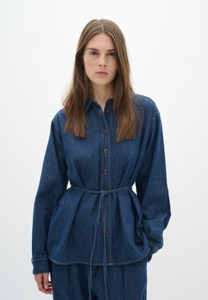 Camicia in denim di colore blu scuro con chiusura a bottoni, maniche lunghe e vita cintrata con cintura, che mette in evidenza un tessuto testurizzato.
