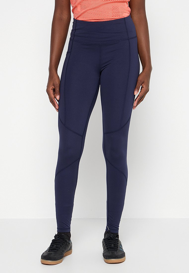 Osoba stojąca z rękami na bokach, ubrana w granatowe legginsy o wysokim stanie, czarne sneakersy i pomarańczową koszulkę sportową.