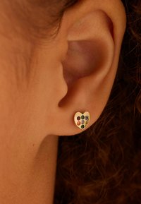 Boucle d'oreille en forme de cœur en or avec des pierres précieuses rondes et colorées, comprenant du vert, du violet, de l'orange et du bleu, se détachant sur un fond texturé.