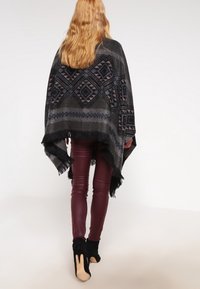 Černá a šedá vzorovaná ponča s třásněmi, nošená přes vínové legíny a černé kotníkové boty. Disponuje geometrickými vzory a jemnou strukturou.