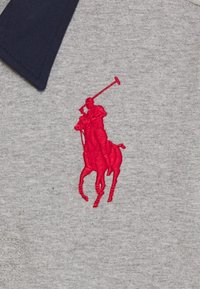 Červená vyšitá ikona hráče póla na koni na šedé látce pod tmavě modrým límcem košile.