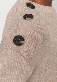 Krémový pletený svetr s texturovaným povrchem, který má čtyři tmavé knoflíky na rameni a přiléhavý střih.