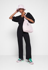 Combinaison noire à manches larges, associée à un chapeau seau rose clair et un sac fourre-tout assorti. Les chaussures ont un design multicolore.