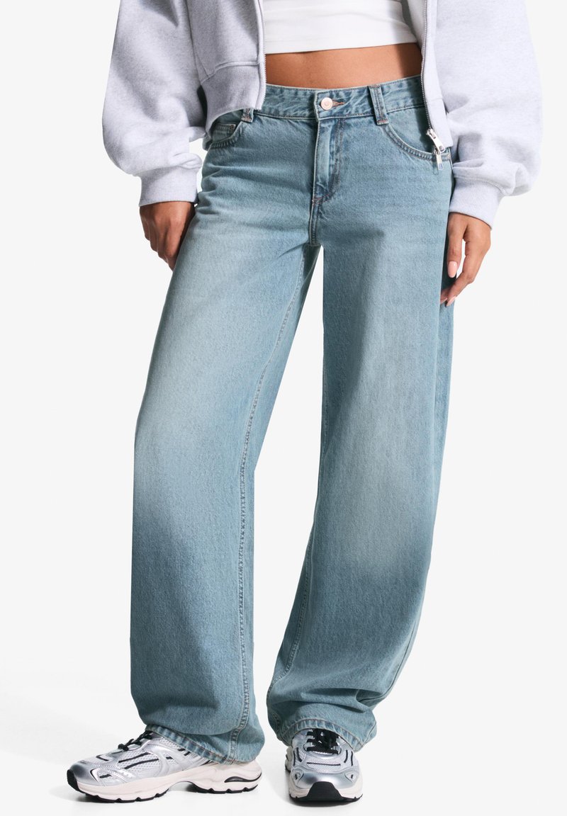 Blugi lejera de culoare albastru deschis, din denim, cu talie înaltă, cusături vizibile și o ușoară decolorare. Purtați cu adidași argintii.