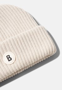 Beżowa, ribbowana czapka beanie z podwiniętym brzegiem i okrągłą naszywką z literą "B" w kolorze czarnym. Gładka struktura i miękki wygląd.