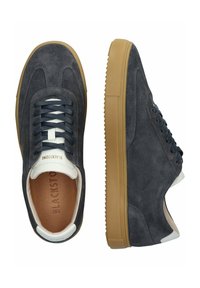 Zapatillas de ante gris con una lengüeta de cuero blanco, cordones azul marino y una suela de goma marrón que presenta un patrón de agarre texturizado.