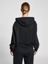 Person mit blondem Haar im Dutt, die einen einfachen schwarzen Hoodie und schwarze Hosen trägt, von hinten vor einem neutral grauen Hintergrund gezeigt.