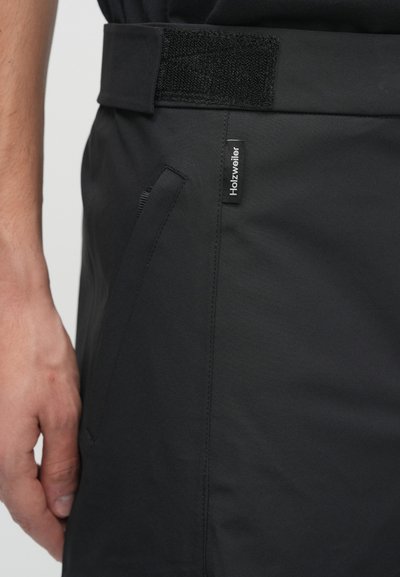 Juodi šortai, pagaminti iš lygios medžiagos, turi Velcro juosmenį, šoninį kišenę ir logotipo etiketę kairėje pusėje.