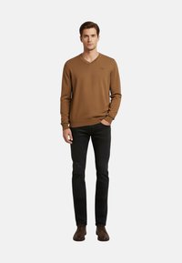 Maglione marrone a scollo a V in tessuto lavorato a maglia, modello aderente, indossato con jeans neri e scarpe marroni; presenta un piccolo dettaglio con logo sul petto.