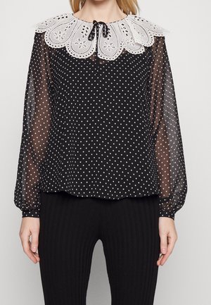 Kobieta w czarnej, przezroczystej bluzce z długim rękawem w białe kropki, z dużym białym, koronkowym kołnierzem o falistym brzegu, zawiązanym czarną wstążką.