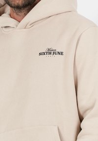 Sudadera con capucha beige hecha de una tela suave, con un pequeño logotipo negro en el pecho que dice "Maison SIXTH JUNE PARIS."