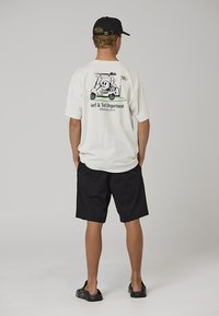Hvid oversized t-shirt med en baggrafik af en tegneseriefigur på en golfvogn, kombineret med sorte shorts og sorte sandaler.