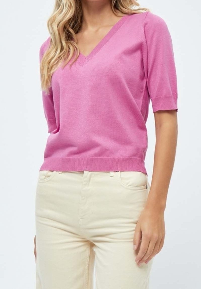 Femme portant un pull rose à manches courtes avec col en V et un pantalon beige clair taille haute, vue du cou jusqu'aux hanches sur un fond uni.