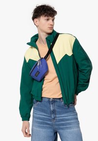 Giacca a vento verde e gialla con polsini elastici, indossata sopra una camicia pesca, abbinata a una borsa pochette blu e jeans azzurri.