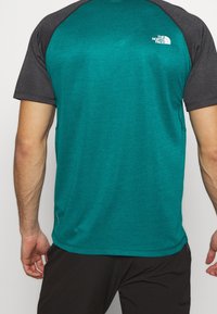 Koszulka sportowa z krótkim rękawem, o turkusowym korpusie i czarnych rękawach raglan, z teksturowanym wzorem i małym białym logo na górnej części pleców.