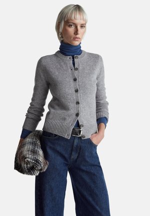 Šedý pletený kardigan s žebrovaným lemem a zapínáním na knoflíky, vrstvený přes modrý rolák, doplněný tmavě modrými džíny s širokými nohavicemi.