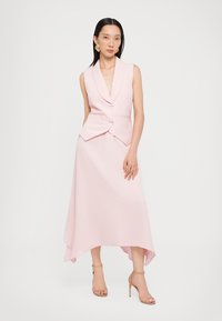 Abito smanicato rosa con silhouette sartoriale, revers affilati e orlo asimmetrico. Presenta due bottoni argentati frontali.