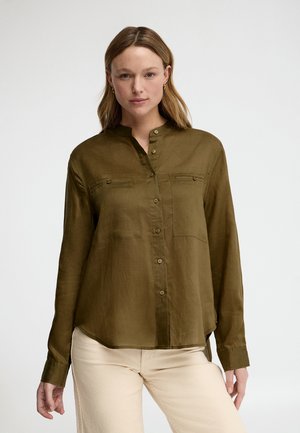 Woman wearing olive green button-up shirt with chest pockets and cream pants, standing against a plain light background.