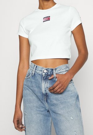 Nainen, joka on pukeutunut valkoiseen lyhythihaiseen Tommy Jeans -t-paitaan ja korkeavyötäröisiin vaaleansinsisiin farkkuihin, yksi käsi taskussa.