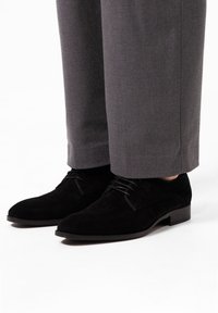 Zwarte suede salonschoenen met een ronde neus, vetersluiting en een lage blokhak, gecombineerd met grijze maatwerkbroek.