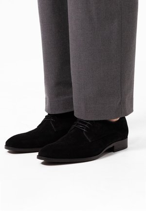 Black suede dress shoes with a rounded toe, lace-up design, and a low block heel, paired with gray tailored trousers.